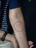 Tatuagem minimalista feita em linha fina (fineline) na região do antebraço, representando o desenho de um coração partiido, entrelaçado com o simbolo do infinito e com a frase "eu só quero encarar o mar e as flores da primavera".