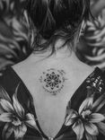 Tatuagem minimalista feita em linha fina (fineline) e pontilhismo (dorwork) nas costas, na região da coluna vertebral, representando uma flor mandala e a frase "rir é um ato de ristência", em volta; de forma circular.