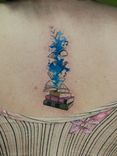 Tatuagem minimalista feita em linha fina (fineline), colorida, estilo aquarela (watercolor) representando uma pilha de livros, "explodindo simbolos e cor azul"m simbolozando os conhecimentos absorvidos.