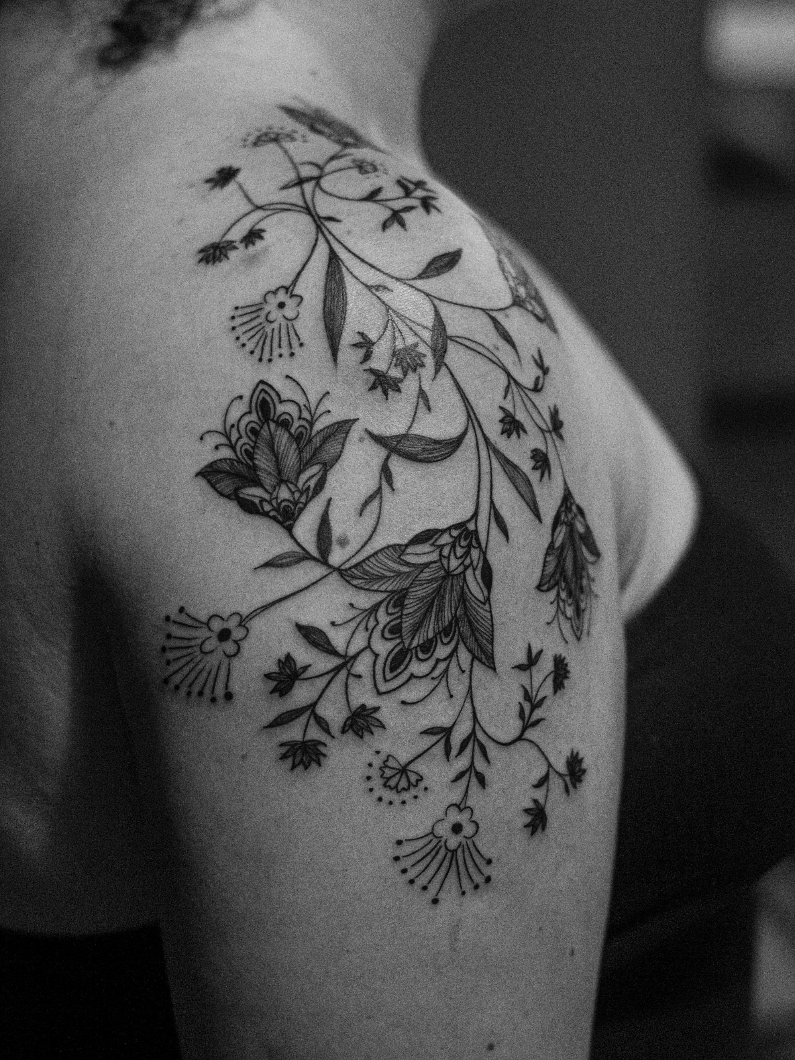 Tatuagem ornamental feita em linha fina (fineline), na região do ombro e braço, somente em preto (blackwork), representando ornamentos e flores mandalas.