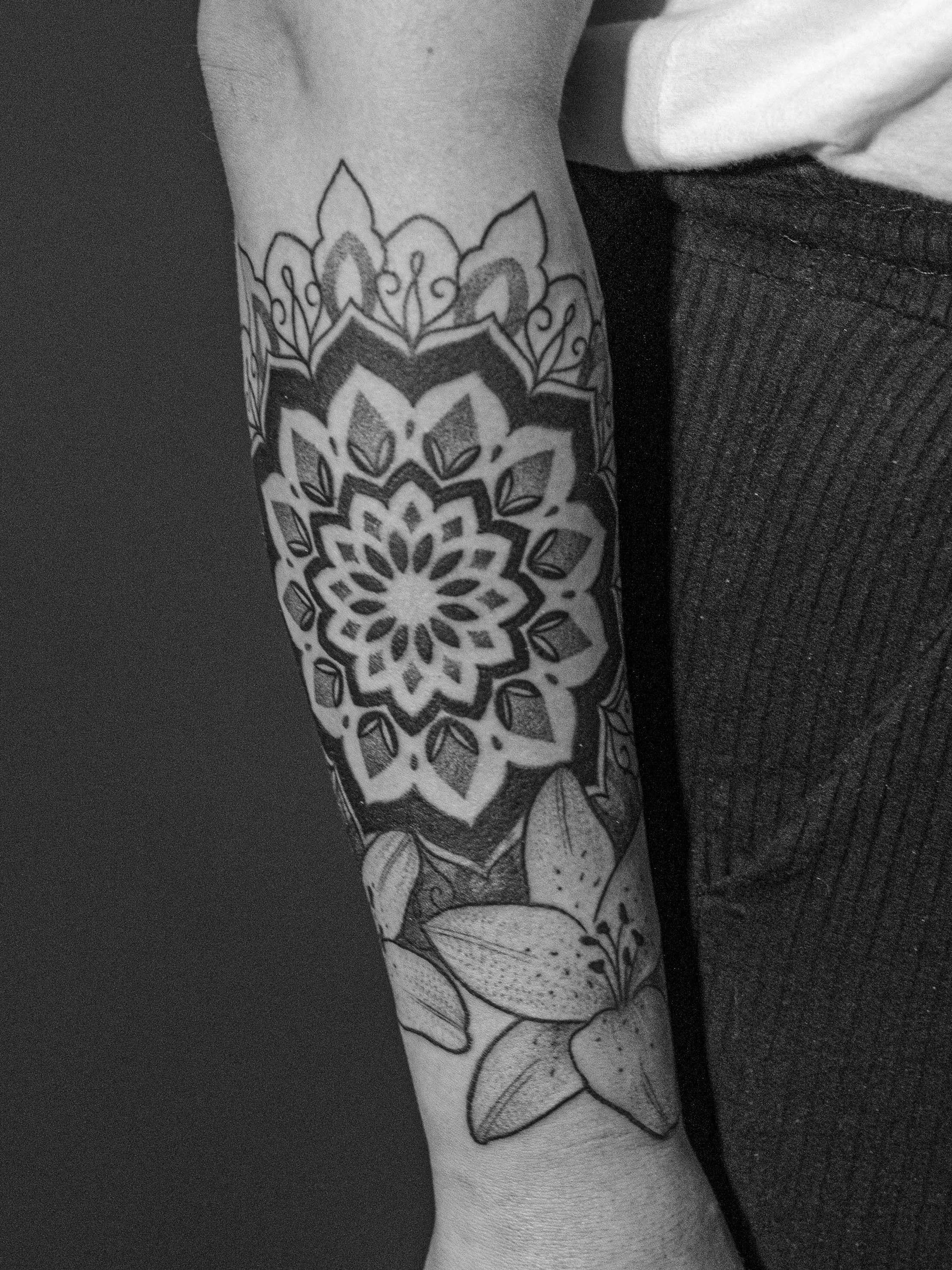 Tatuagem feita em pontilhismo (dotwork) na região do antebraço, representando uma flor/mandala e com dois lírios de complemento.