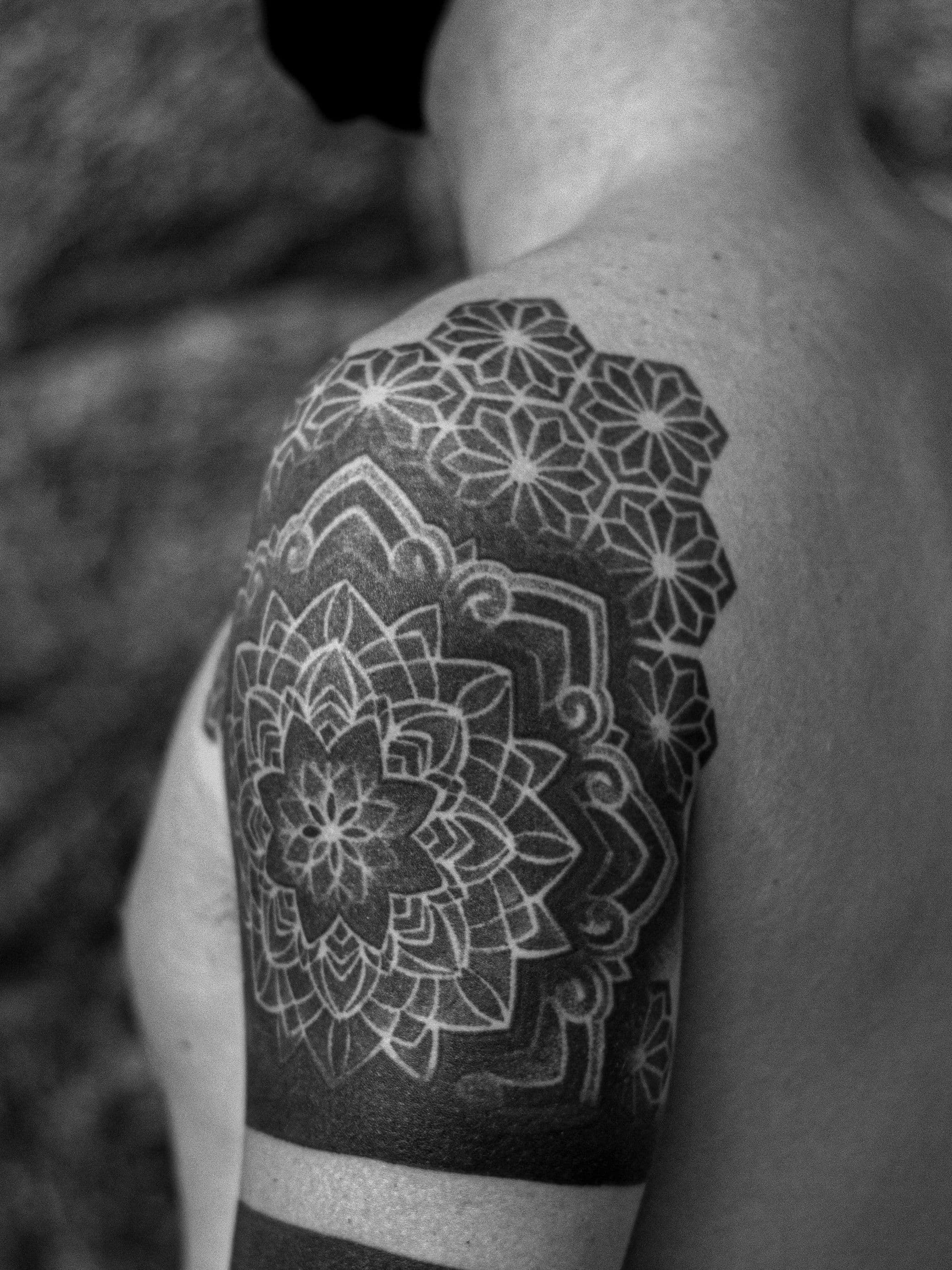 Tatuagem feita em pontilhismo (dotwork) na região do ombro e braço, representando uma flor/mandala e padrões geométricos.