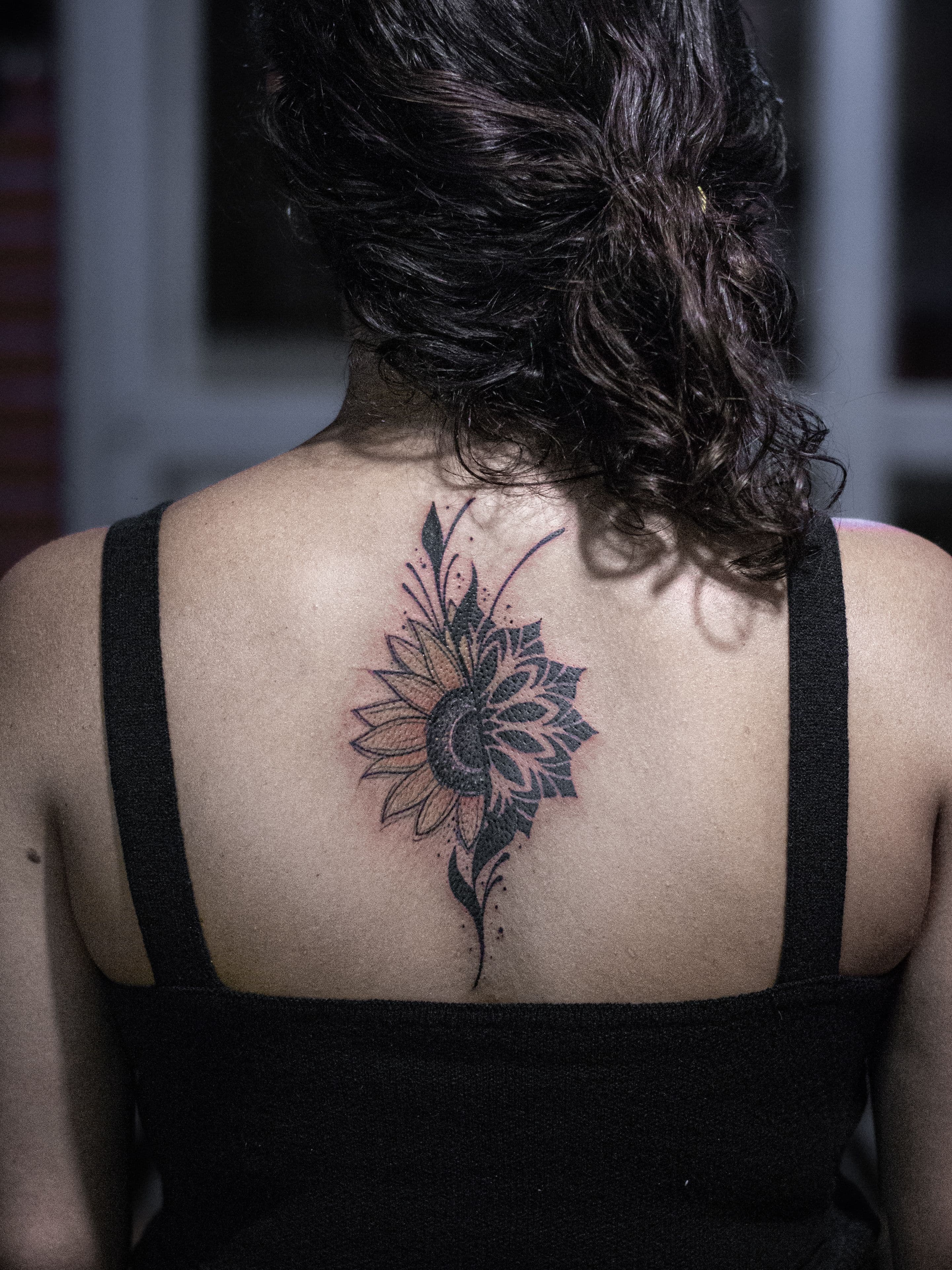 Tatuagem feita em linha fina (fineline), colorida, estilo esboço (sketch), representandO metade de um girasol e metade de uma mandala em pontilhismo (dotwork).