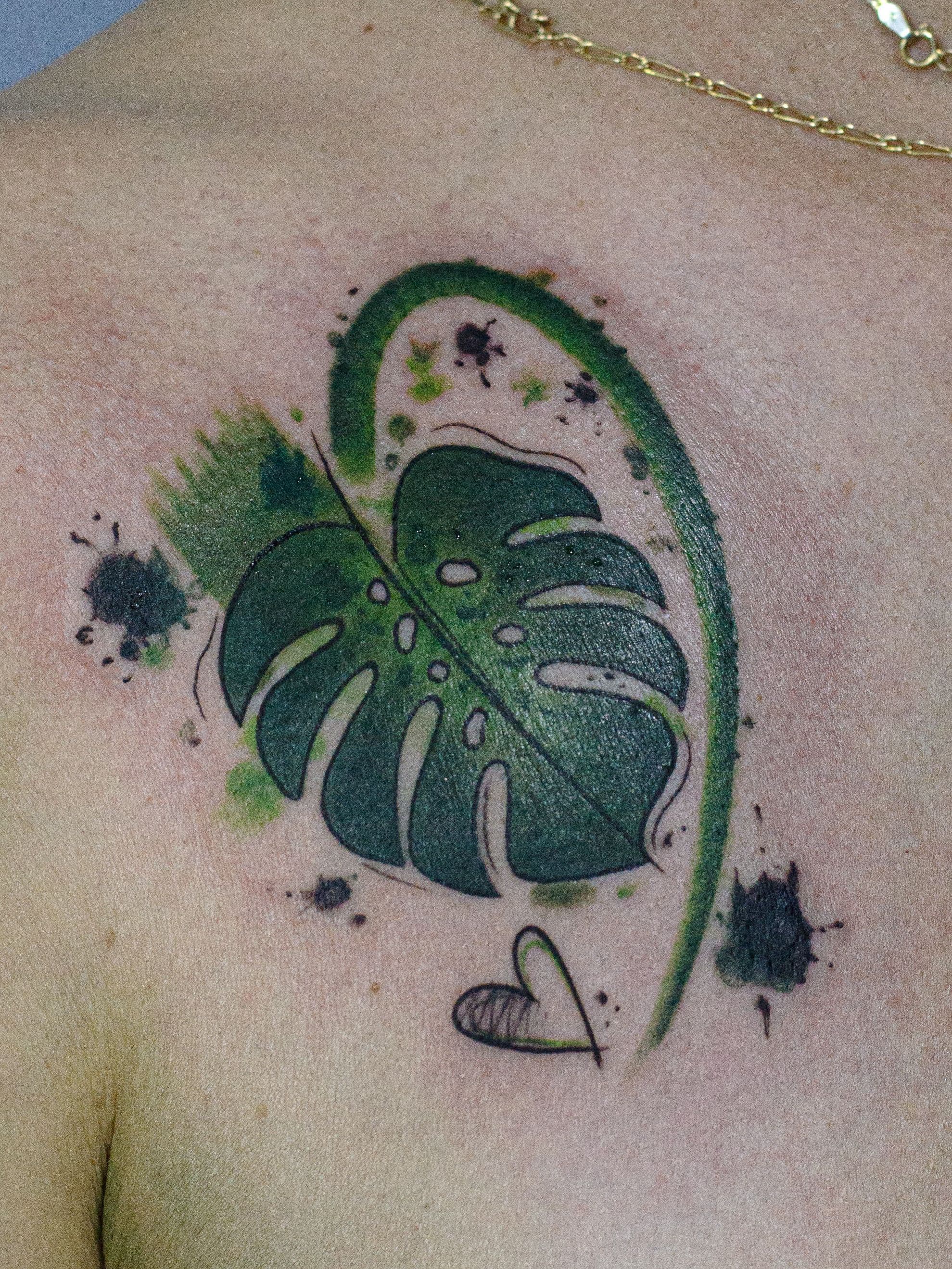 Tatuagem feita em linha fina (fineline), colorida estilo aquarela (watercolor), estilo esboço (sketch), representando uma folha da planta "costela de adão".