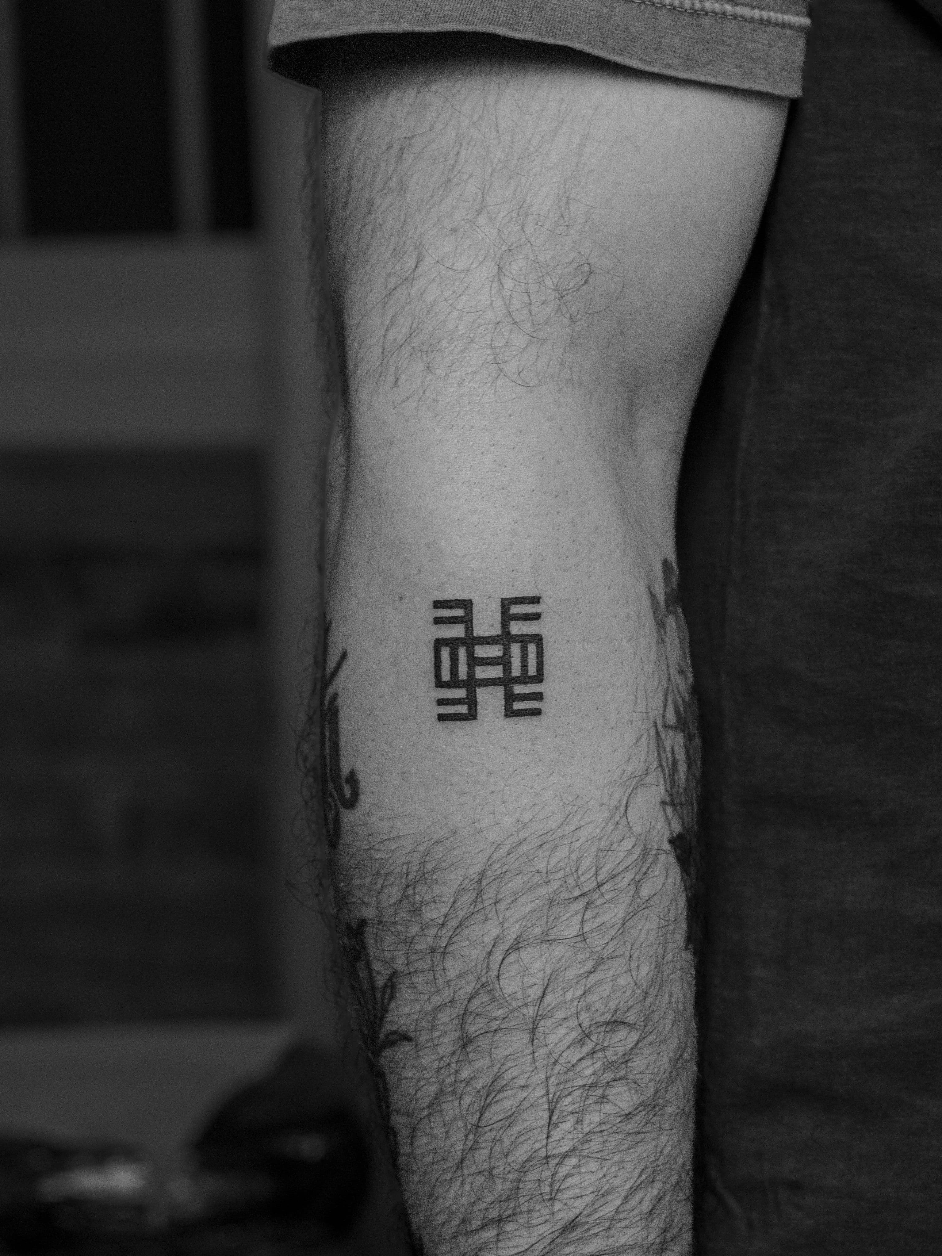 Tatuagem minimalista, someente em preto (blackwork), na região do antebraço, representando o simbolo Adinkra "Nea onnim no sua a, ohu” significa que “aquele que não sabe, pode aprender”.