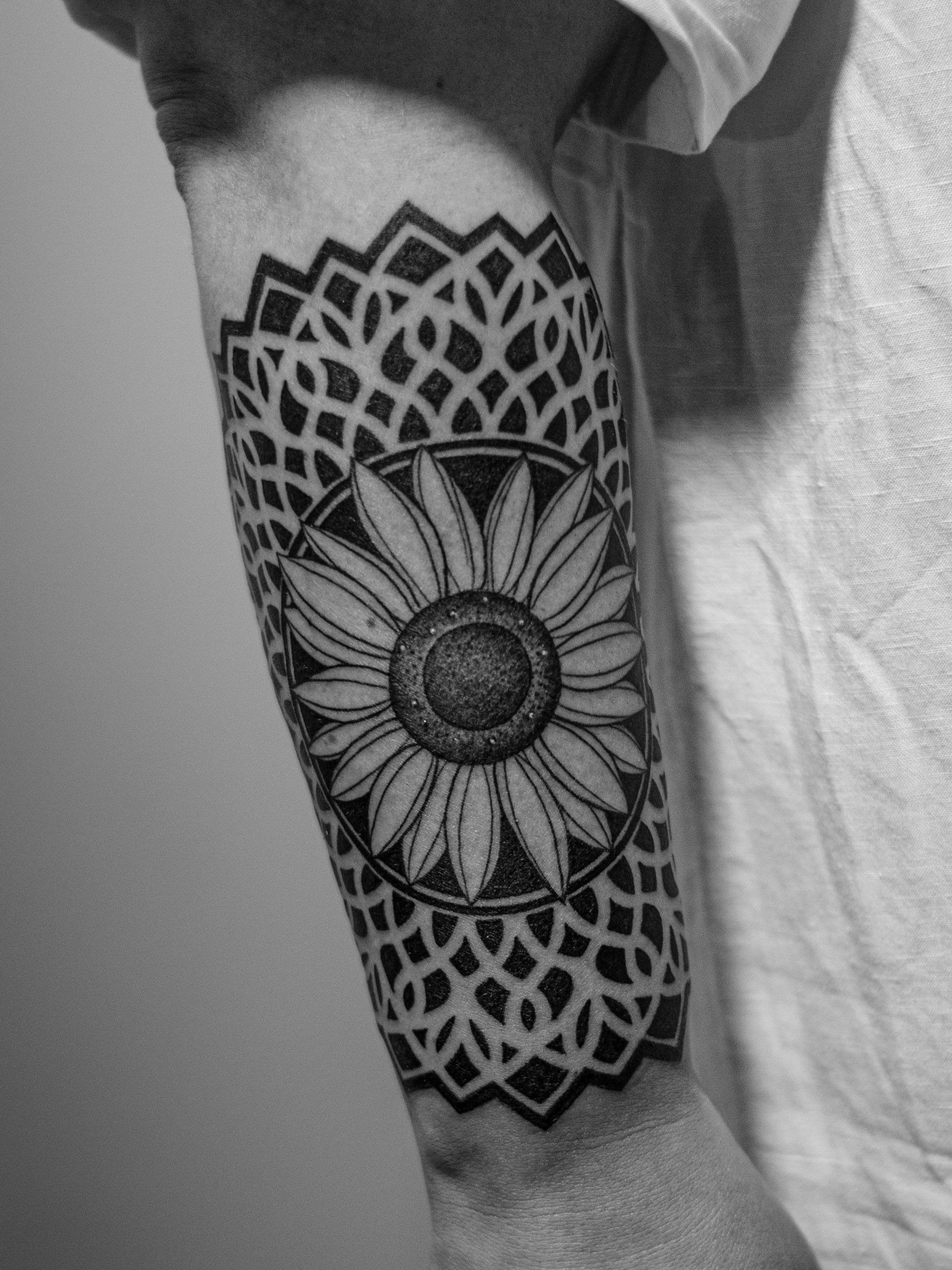 Tatuagem geométrica, feita em pontilhismo (dotwork), somente em preto (blackwork), na região do antebraço, representando uma mandala com um girasol no centro.