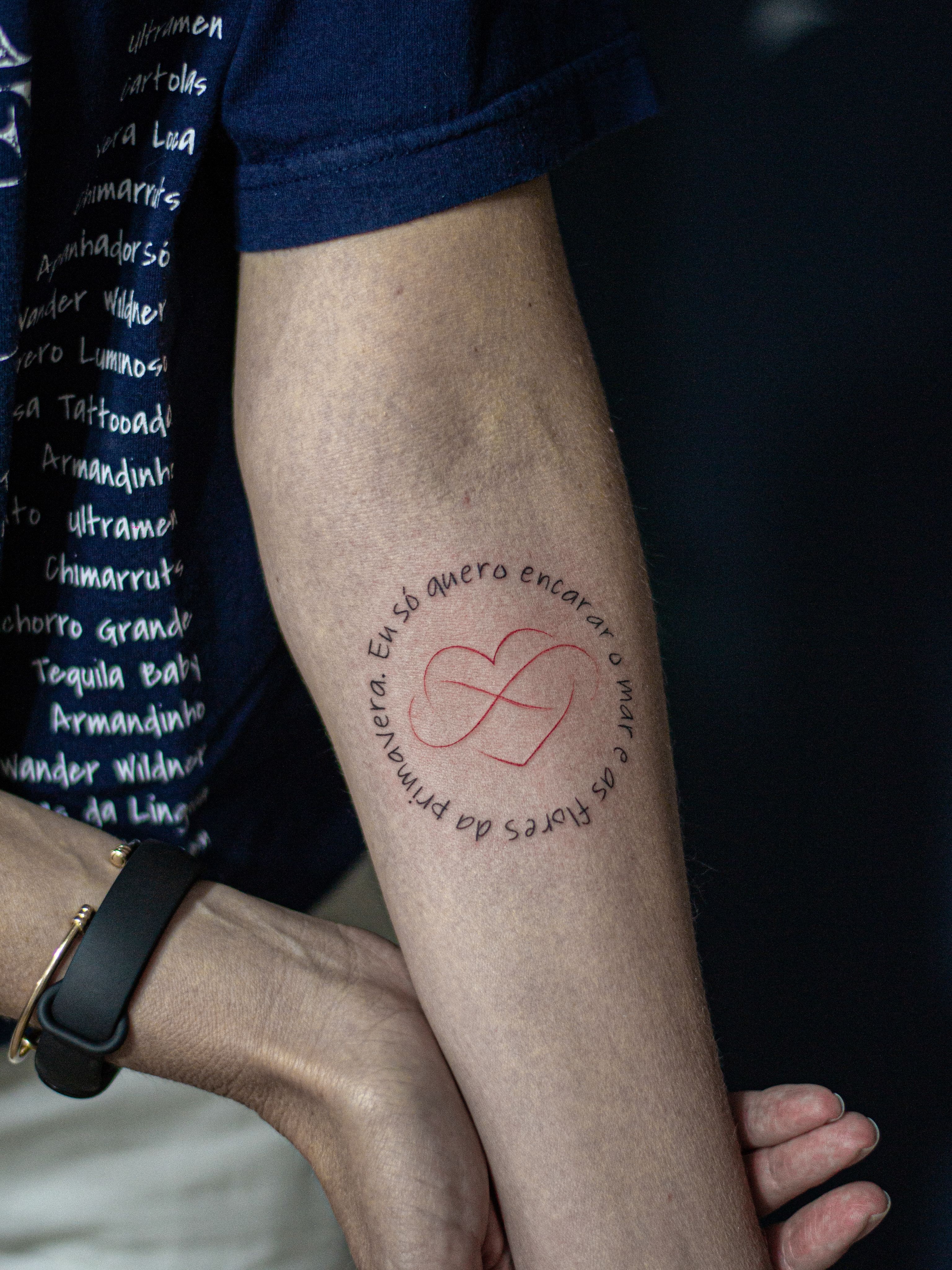 Tatuagem minimalista feita em linha fina (fineline) na região do antebraço, representando o desenho de um coração partiido, entrelaçado com o simbolo do infinito e com a frase "eu só quero encarar o mar e as flores da primavera".