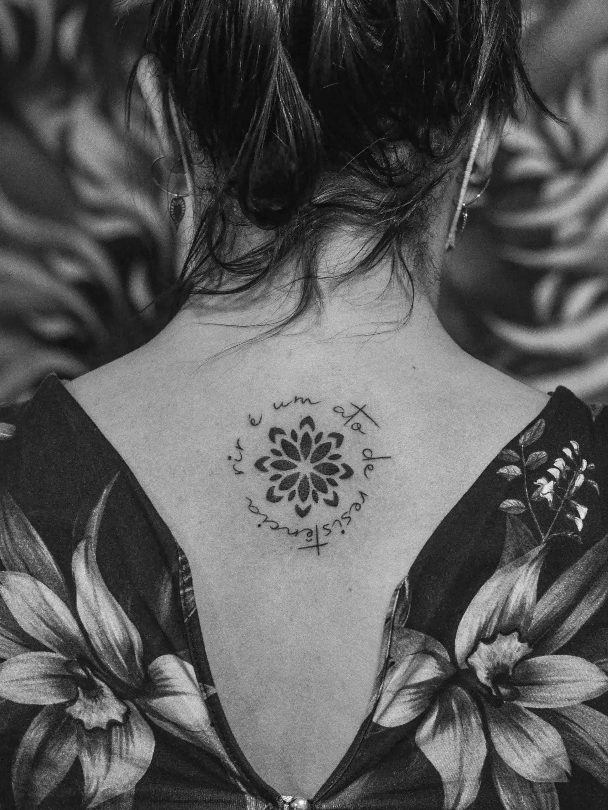 Tatuagem minimalista feita em linha fina (fineline) e pontilhismo (dorwork) nas costas, na região da coluna vertebral, representando uma flor mandala e a frase "rir é um ato de ristência", em volta; de forma circular.