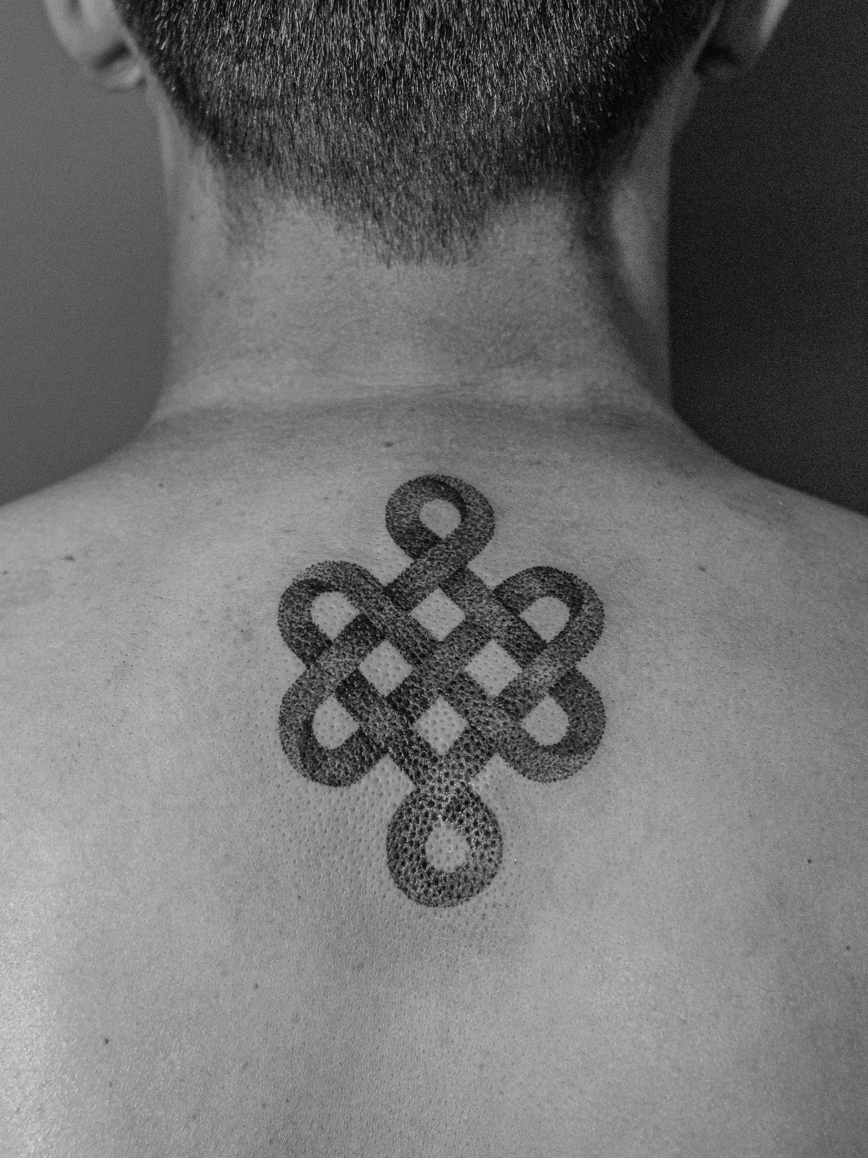 Tatuagem minimalista feita pontilhismo (dorwork) na região da costas, próximo ao pescoço, representando o simbolo budista "nó infinito".