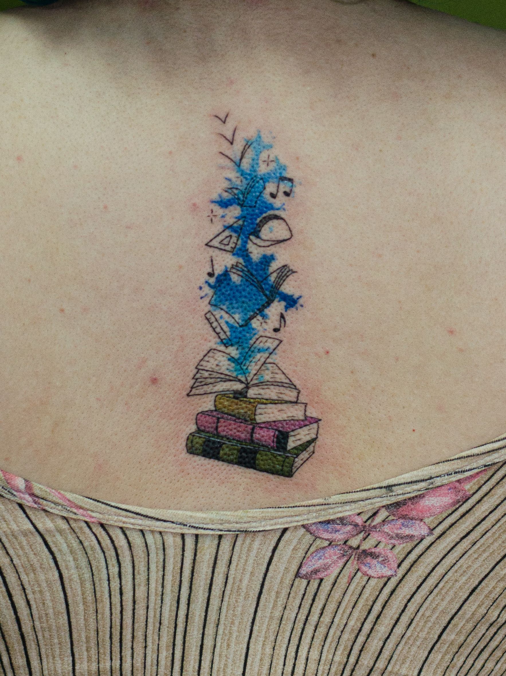 Tatuagem minimalista feita em linha fina (fineline), colorida, estilo aquarela (watercolor) representando uma pilha de livros, "explodindo simbolos e cor azul"m simbolozando os conhecimentos absorvidos.